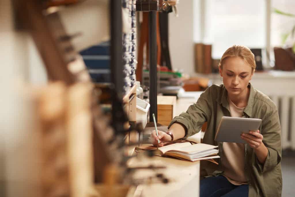 young blond woman sitting at table in craft workshop looking at tablet PC screen and writing down something in notebook